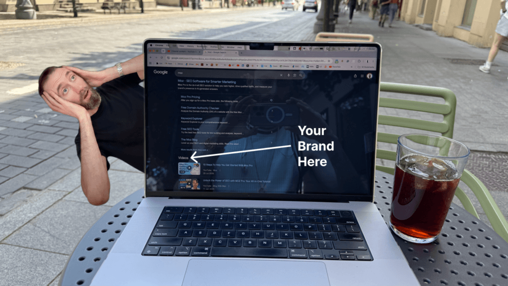 Laptop on an outdoor cafe table showing a Google search for “moz,” with video results under the brand’s homepage and a white arrow labeled “Your Brand Here,” illustrating how competitor brand searches can surface YouTube videos before buyers choose another company.