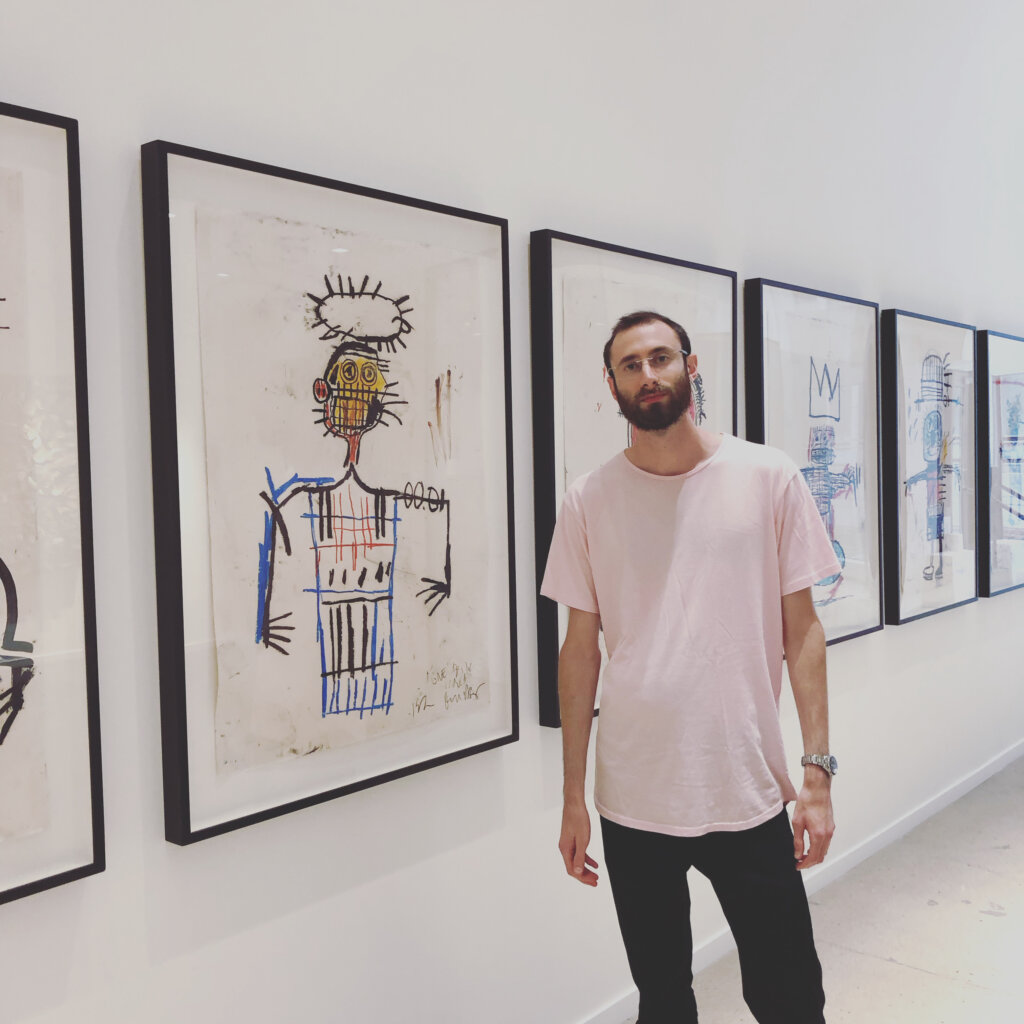 Edward Sturm in a pink shirt at a private gallery, standing with a line of Jean-Michel Basquiat paintings. Photo taken in NYC in 2023.