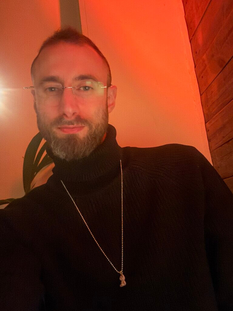 Edward Sturm wearing a bear necklace and cool af turtleneck sweater at a restaurant in Europe in 2025.