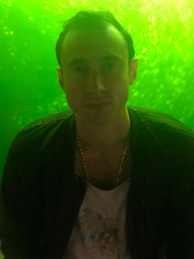 Edward Sturm wearing a gold chain with green bubbled liquid behind him. Photo taken in Belgrade, Serbia in 2021.