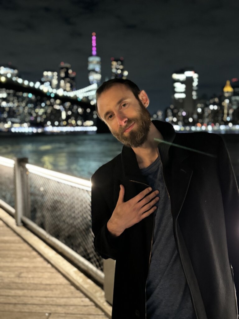 Edward Sturm at night in DUMBO, NYC. The East River, Brooklyn Bridge, and Lower Manhattan behind him. Photo taken in 2023.