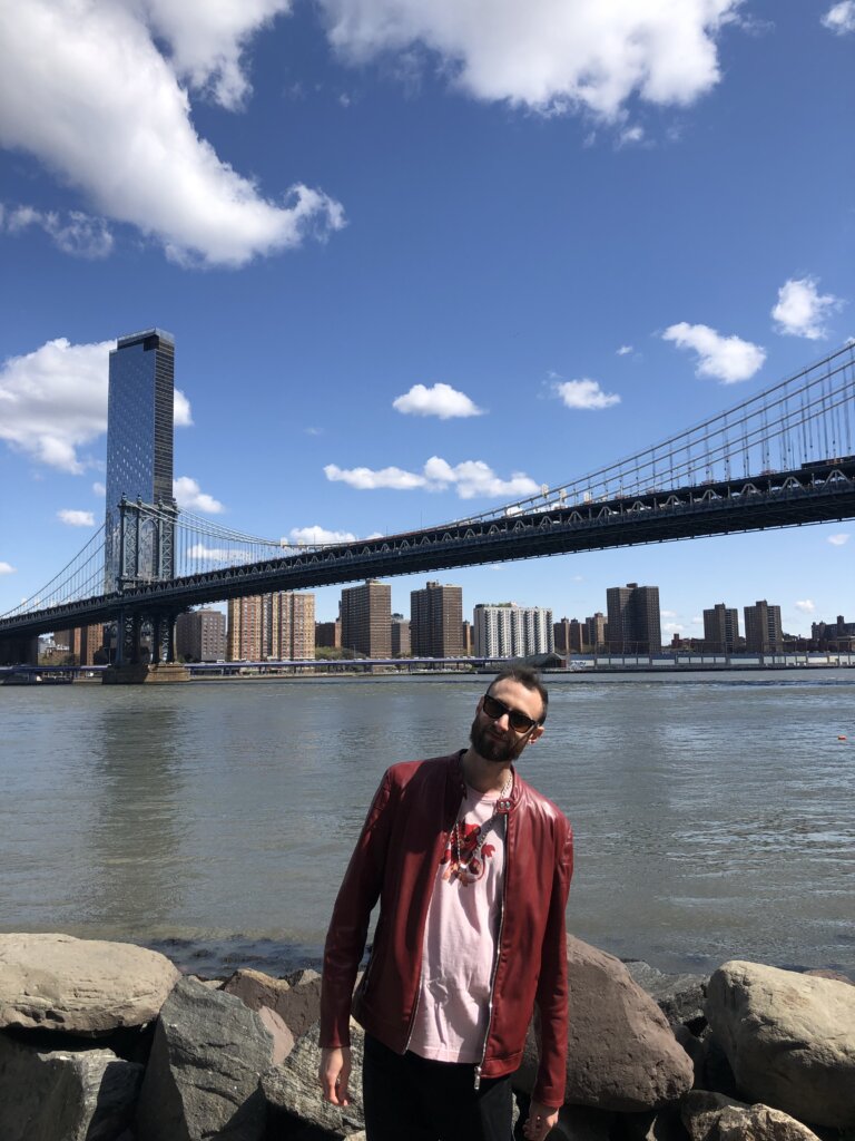 Edward Sturm in a red leather jacket and Matchstick Bear t-shirt in front of the East River and Manhattan Bridge in NYC. Photo taken in 2022.