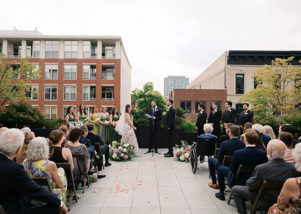 Edward Sturm officiating his brother's rooftop wedding, standing between the bride and groom with bridesmaids in lovely green dresses, groomsmen in handsome black suits, and guests seated on both sides of a petal-strewn aisle, Chicago, 2026.