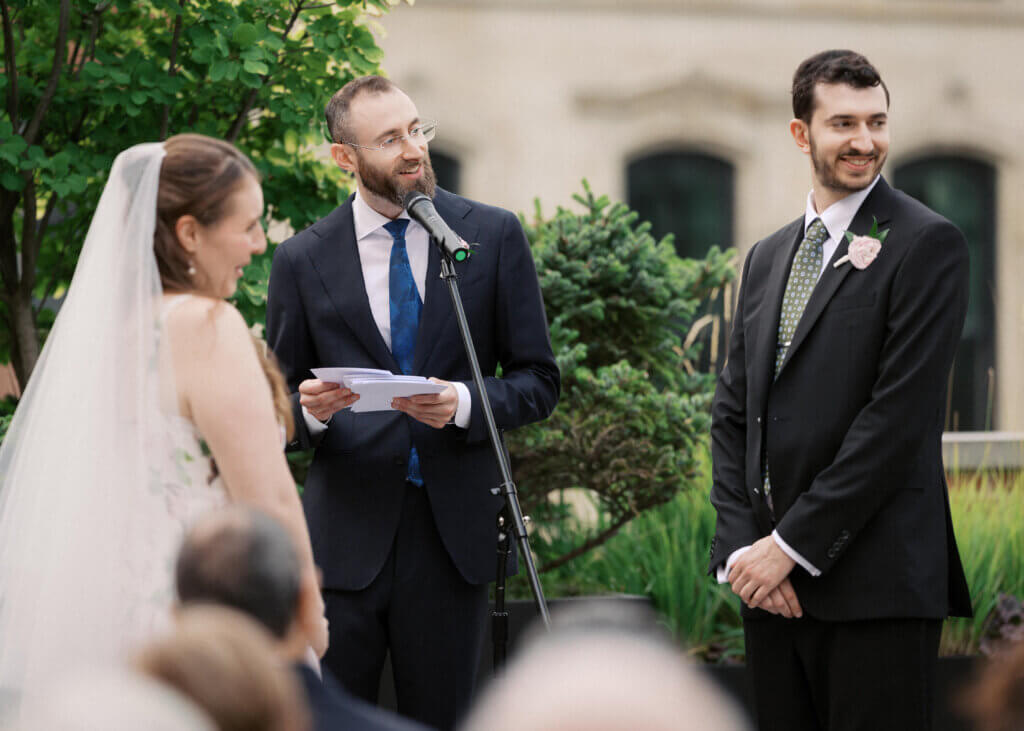 Edward Sturm smiling while officiating his brother's wedding, holding his notes at the microphone as his brother, the groom, smiles beside him and the bride in her veil looks on, Chicago - 2026.