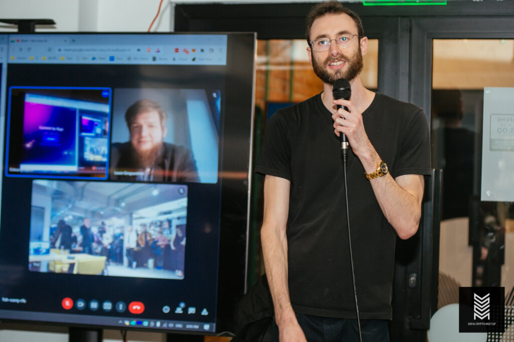 Edward Sturm wearing a gold watch speaking on stage with his cofounder, Leon Grapenthin, at the Sofia, Bulgaria crypto meetup. Edward is speaking about his project, Commit Club. Photo taken in 2022.